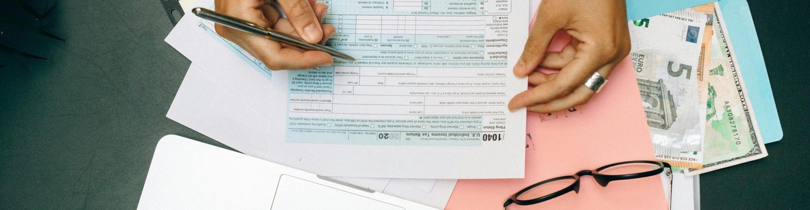 Hands writing on tax documents with laptop, glasses, and currency on desk.